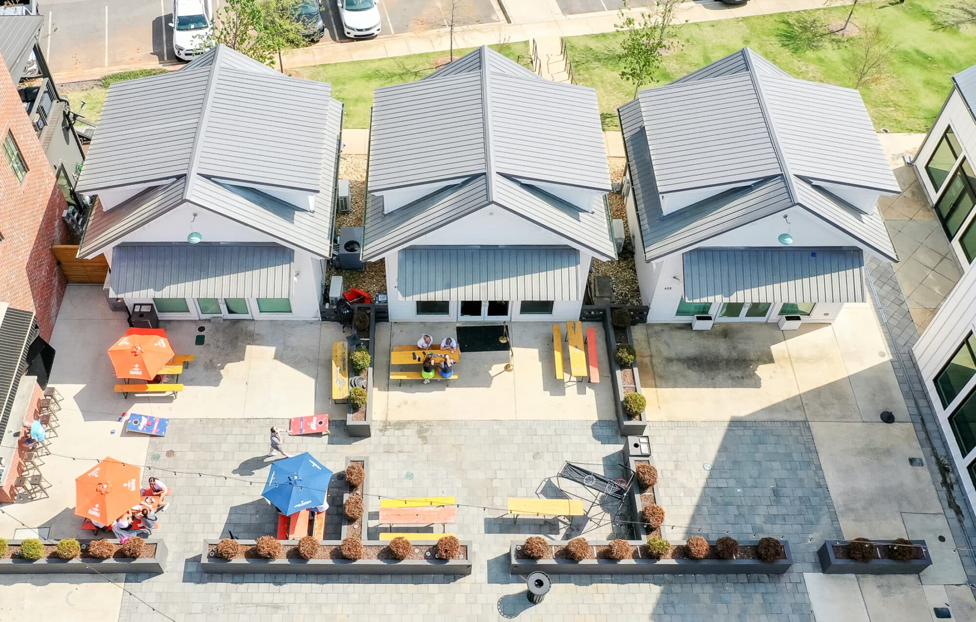An aerial view of three small buildings at Midtown Auburn with gray rooftops surrounded by a public picnic area. The picnic tables are arranged in a square formation with a tree in the center, providing shade for those seated at the tables. The surrounding area is covered with green grass, and there are small bushes and trees dotted around the area.