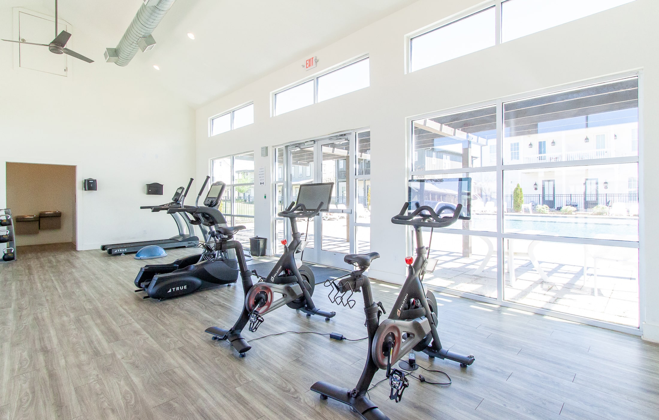 A brightly lit fitness room with large windows overlooking a cityscape, located at Midtown Auburn. The room is equipped with two Peloton cycles and a treadmill, offering a variety of workout options. The Peloton cycles are located at the forefront of the room, with sleek screens displaying fitness metrics and other information. The treadmill is positioned towards the back of the room, with a fan and a water bottle holder attached to it. The room is decorated with motivational quotes and has a mirror spanning the length of one of the walls, allowing gym-goers to check their form. The flooring is made of black rubber tiles, and the ceiling features several industrial-style light fixtures. Overall, the scene suggests a modern and well-equipped fitness facility at Midtown Auburn.